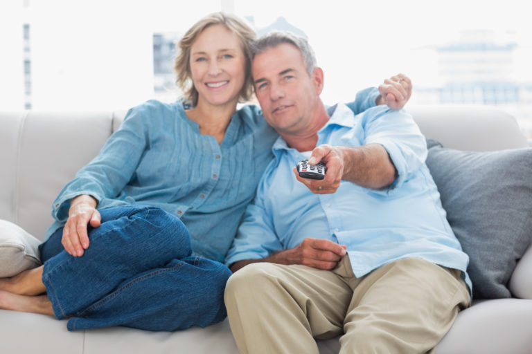 זוג מבוגר שצופה בטלוויזיה מחייכת צוחקת על ספה