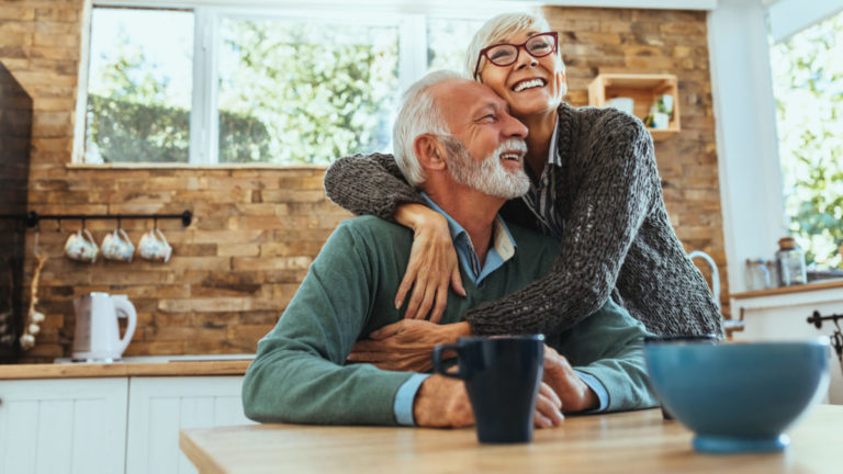 A senior woman hugs her husband in the kitchen.